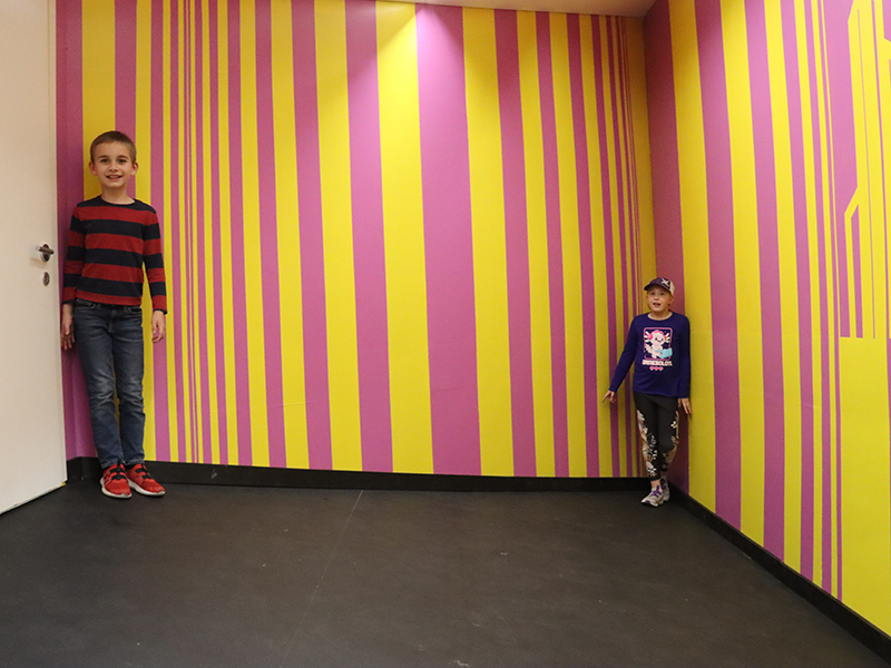 young boy and young girl in the same room but one appears much bigger than the other due to the eye being tricked by depth, perspective, and optical illusions contained within the wallpaper patterns