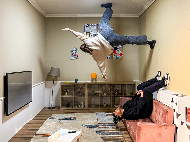 two young adults traversing the zero-gravity room, young man sitting upside-down on couch with feet pressed against the wall and the young woman above him walking upside-down with one foot on the ceiling and the other outstretched and touching the adjoining wall