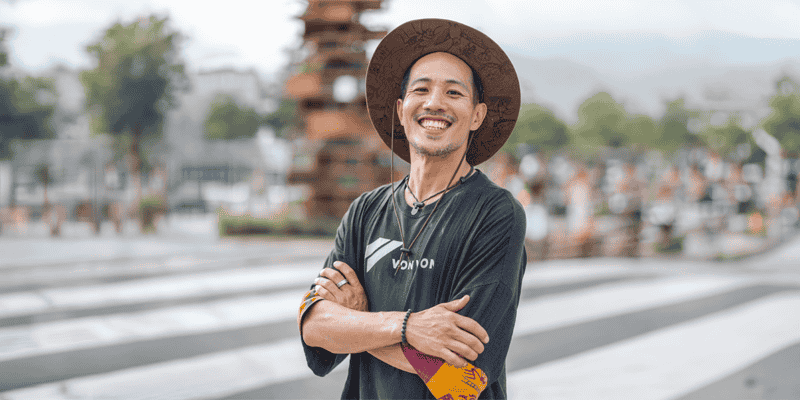 Portrait‑style photograph of a person standing outdoors with arms crossed, wearing a wide‑brimmed hat, a dark long‑sleeve shirt, and patterned wristbands. The background shows an urban setting with a crosswalk, trees, and stacked sculptural structures softly out of focus.