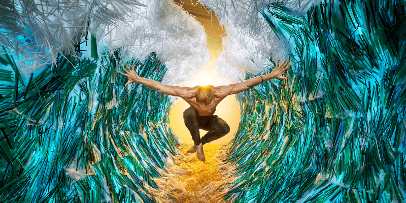 Artistic scene of a person captured mid‑jump between two towering walls of teal and white plastic strips, arranged to resemble crashing waves. A warm golden light shines through the opening behind the figure, creating strong contrast with the textured plastic surroundings.