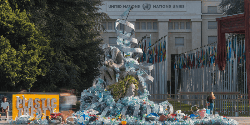Large outdoor sculpture made from discarded plastic waste displayed in front of the United Nations building. The installation features stacked human‑like forms and flowing plastic elements rising upward, surrounded by scattered plastic debris at ground level.