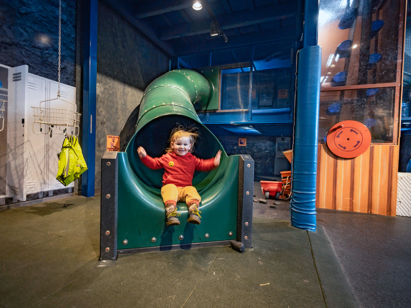 Un jeune enfant vêtu d'un sweat-shirt rouge et d'un pantalon jaune glisse hors de la bouche d'un grand toboggan vert en spirale à l'intérieur d'une aire de jeux/exposition couverte. L'enfant a les bras levés et sourit. L'espace autour du toboggan est délimité par une structure métallique bleue, des poteaux de sécurité rembourrés et un sol en caoutchouc foncé ; des casiers et des gilets haute visibilité suspendus sont visibles sur la gauche.