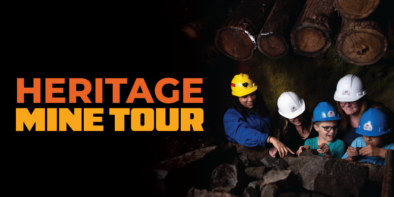 Promotional graphic with bold orange text reading “Heritage Mine Tour” on a dark background. A group of visitors wearing hard hats stands inside a mine, leaning over rocks and examining the display beneath stacked timber supports.
