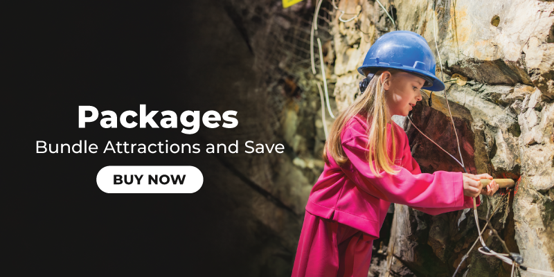 Wide banner with a dark gradient panel on the left and a photo on the right. Left side text reads “Packages” and “Bundle Attractions and Save,” with a rounded “BUY NOW” button underneath. On the right, a child wearing a blue hard hat and bright pink jacket uses a small hammer against a rocky wall, suggesting a mining or geology activity.