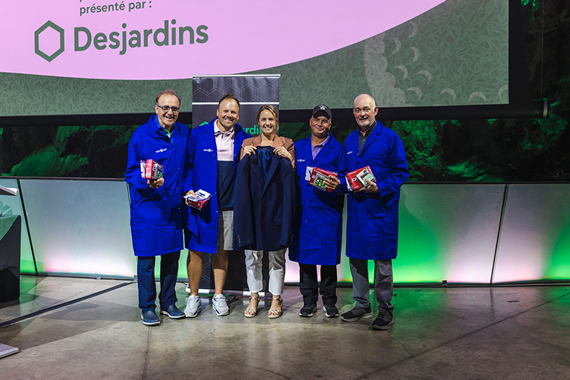 Team Lundrigan, winners of the 2025 Bluecoat Open charity golf tournament hosted by Science North, stand on stage during an awards presentation. Four team members wearing blue Science North lab coats pose alongside a presenter holding a blue blazer. Several team members hold prize boxes. A large screen displaying the Desjardins logo, an event sponsor, is visible above the group.